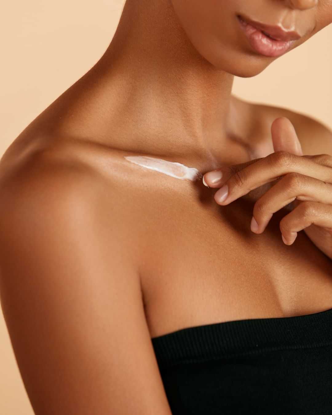 Woman applying lotion to shoulder and collarbone, wearing a black strapless top.